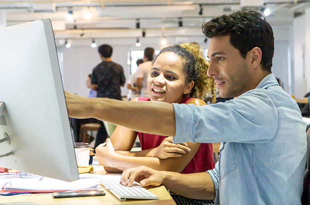 Two coworkers at a desk look at a computer, with one pointing at the screen while explaining something.