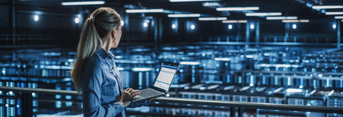 A woman stands in a large, high-tech data center, holding a laptop and looking over rows of illuminated server racks.