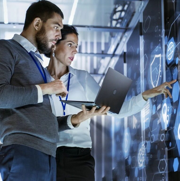 Two professionals with badges collaborate in a tech environment, one holding a laptop while the other points at a digital interface on a large screen.