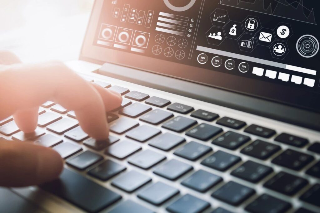 Close-up of a person typing on a laptop keyboard with one hand, while a screen above shows a digital dashboard with charts, icons, and data visualizations.
